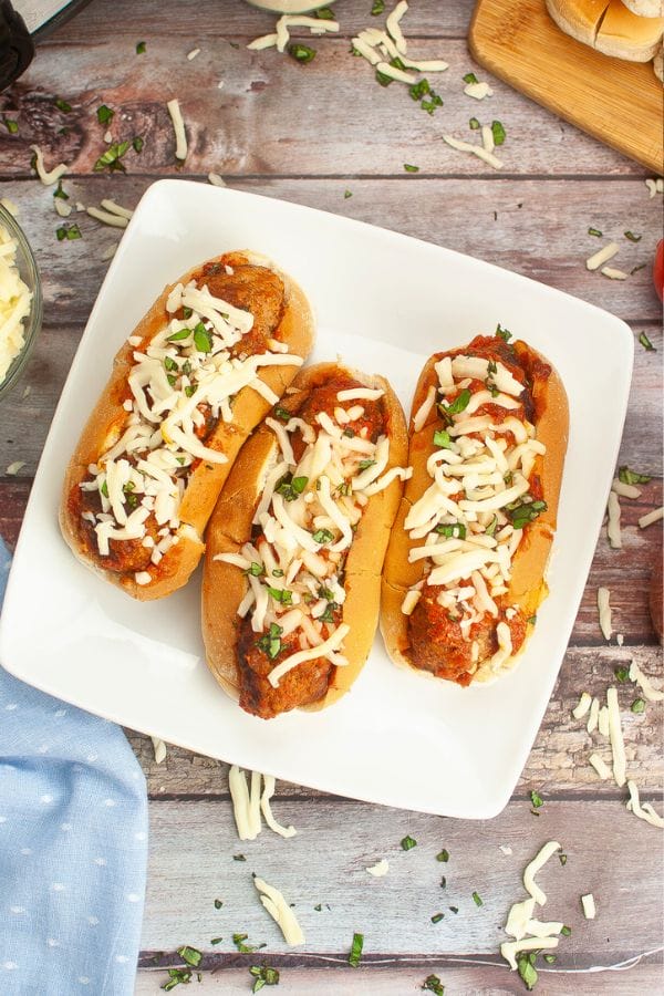 Three meatball subs topped with melted cheese and chopped herbs sit invitingly on a white plate, perfect for a kid-friendly dinner, and placed on a rustic wooden table.