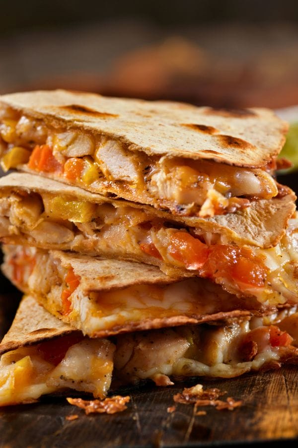 Stack of cheesy chicken quesadillas with visible diced tomatoes and peppers, cut into triangles and placed on a wooden surface, perfect for a kid-friendly dinner.