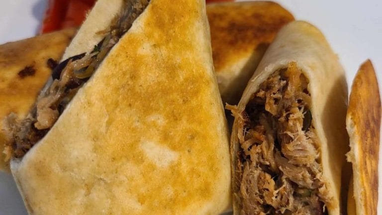 Close-up of toasted wraps stuffed with shredded meat and vegetables, reminiscent of easy pork burritos. A few red slices, possibly peppers, are visible in the background.