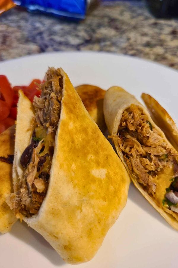 A close-up of an Easy Pork Burrito filled with shredded meat, black beans, and greens. It sits invitingly on a white plate, accompanied by vibrant diced tomatoes on the side.