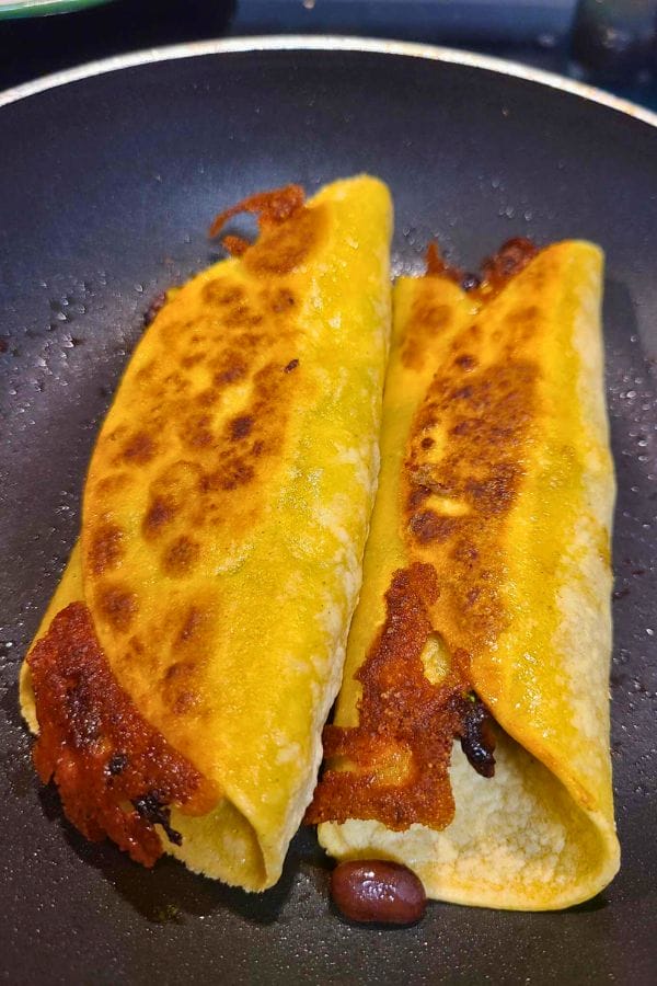 Two crispy, golden-brown tacos filled with melted cheese and beans simmer alongside easy pork burritos in a sizzling frying pan.