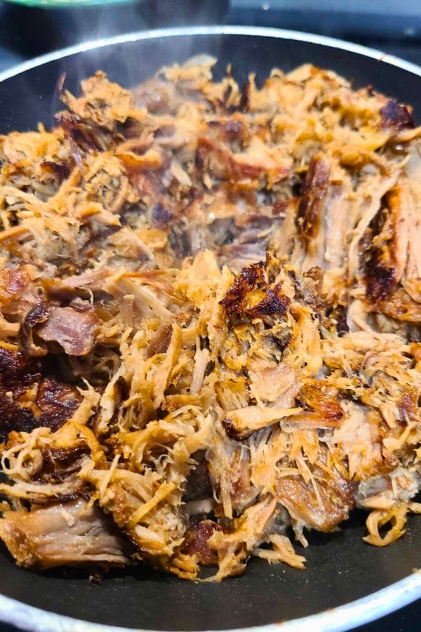 Close-up of shredded, cooked meat in a pan, with some pieces showing a crispy texture—perfect for easy pork burritos. Steam rises from the dish, indicating it is hot and ready to savor.