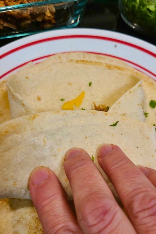 Close-up of a hand holding a folded tortilla on a plate, with visible melted cheese and herbs, making these easy pork burritos irresistible.