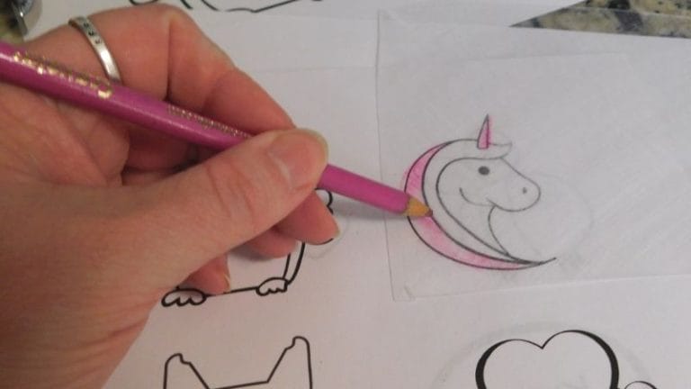A hand with a ring holds a pink pencil, sketching a smiling unicorn head with a pink mane on tracing paper amid an array of free Shrinky Dink templates.