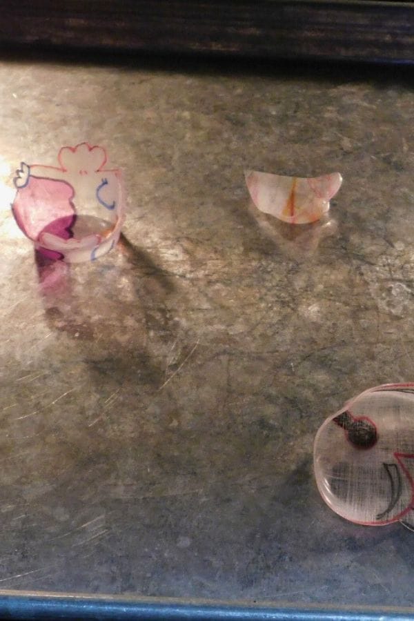 Three transparent Shrinky Dinks, crafted using free Shrinky Dink templates and showcasing simple colored designs, rest on a metallic surface.