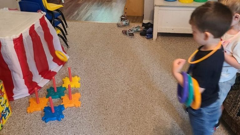 Two children are enjoying a ring toss game indoors, a classic reminiscent of circus activities. One child expertly tosses rings toward colorful pegs while shoes rest near the door, adding a homey backdrop to their playful challenge.