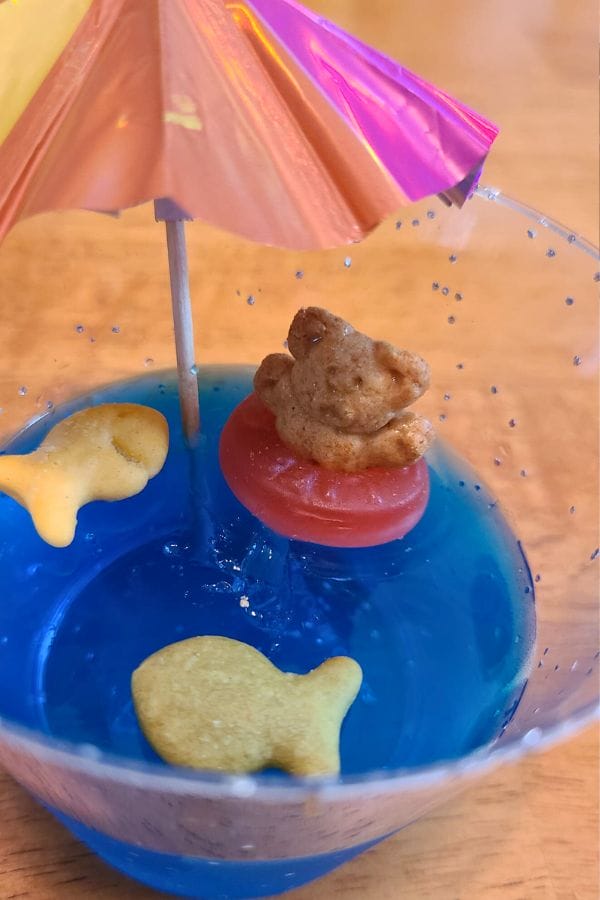 A whimsical blue jelly dessert features a teddy bear cracker lounging on a red candy float beneath a colorful cocktail umbrella, reminiscent of circus activities. Two fish-shaped crackers join the playful scene, adding to the festive allure.