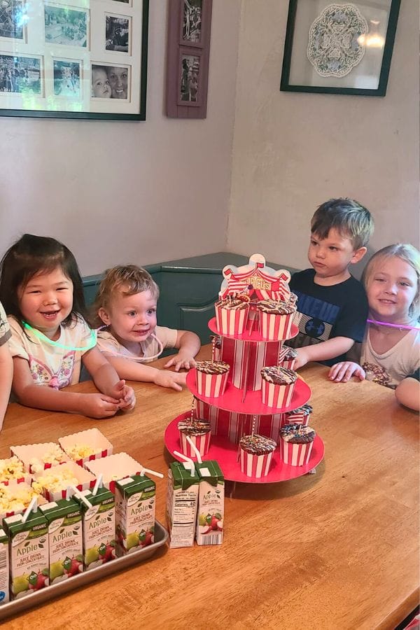 Four children sit at a table with a tiered stand of cupcakes and apple juice boxes, reminiscing about their recent circus activities with excitement sparkling in their eyes.