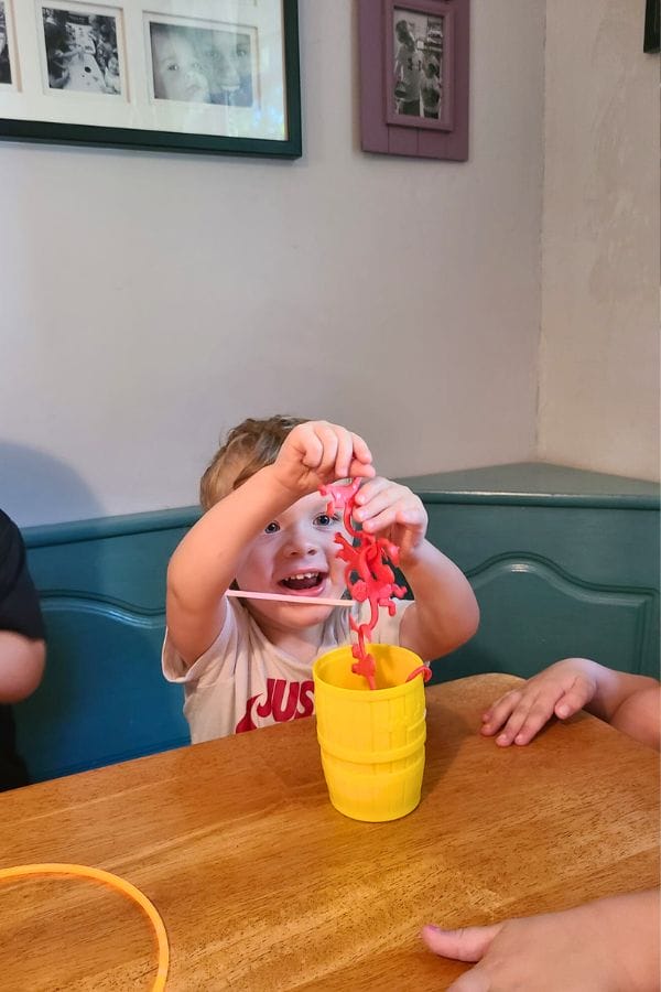 A child excitedly plays with a barrel of monkeys, as if performing circus activities, holding a chain of red monkeys over a yellow cup on a wooden table.