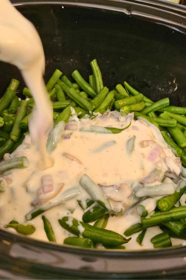 A creamy sauce is elegantly poured over fresh green beans and chopped onions in a slow cooker, creating the perfect base for a delightful Green Bean Chicken Casserole.