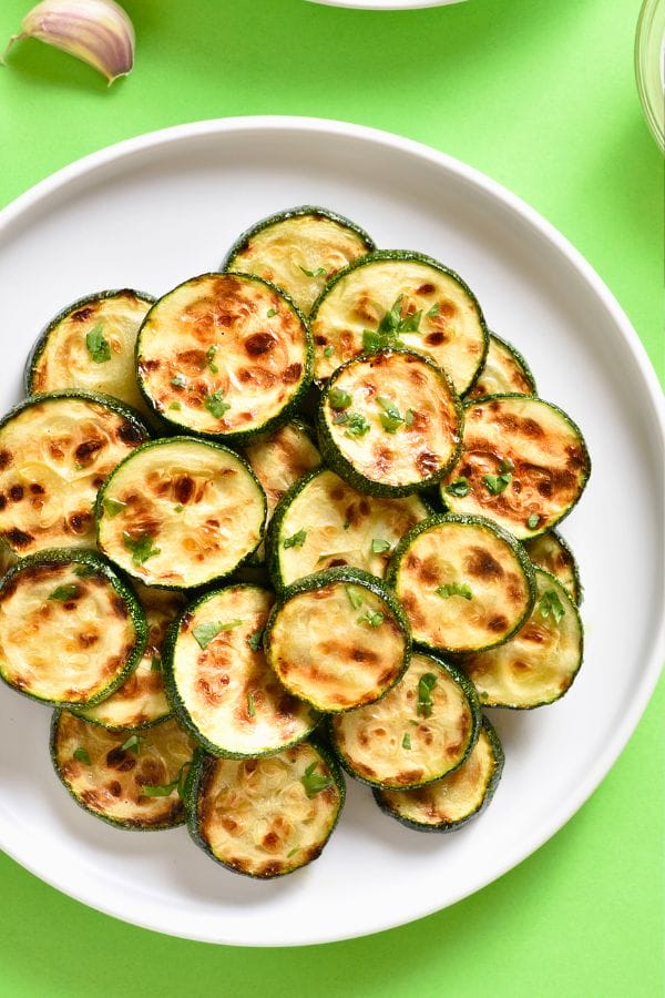 A plate of grilled zucchini slices with herbs sits on a green background, showcasing the fresh bounty from those who choose to grow zucchini vertically.