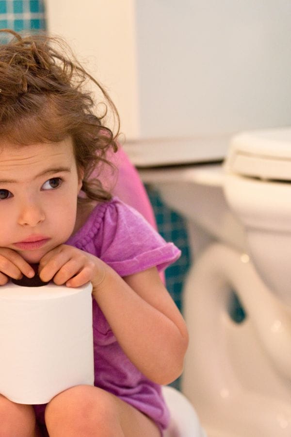 A young child with curly hair, learning how to potty train, sits on a toilet holding a roll of toilet paper while wearing a purple shirt. The bathroom features blue tiles, creating a calm environment for this important milestone.