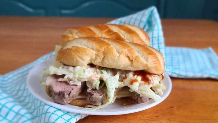 Two sandwiches featuring leftover pork tenderloin, coleslaw, and sauce sit on a plate. They rest on a wooden table adorned with a blue and white checkered cloth in the background.