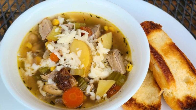 A hearty bowl of soup brimming with sliced vegetables, tender leftover pork tenderloin, and grated cheese, served alongside two pieces of toasted bread on a crisp white plate.