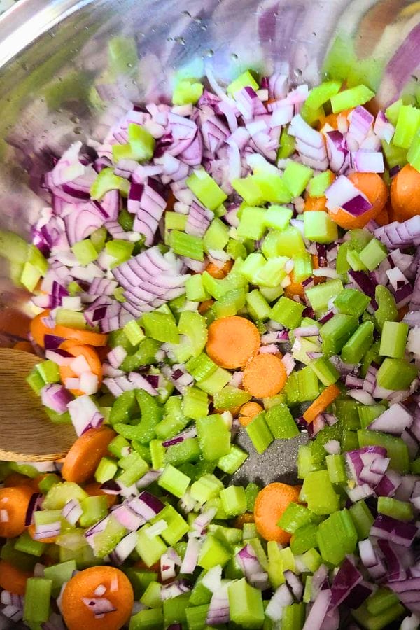Chopped red onions, celery, and carrots simmer alongside leftover pork tenderloin in a stainless steel pot, stirred gently with a wooden spoon.