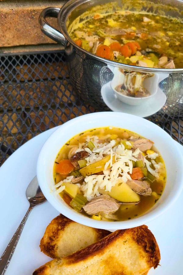 A bowl of soup featuring leftover pork tenderloin, rice, and vegetables is topped with shredded cheese on a white plate accompanied by two pieces of toasted bread. In the background, a pot brims with more of this comforting tenderloin soup.