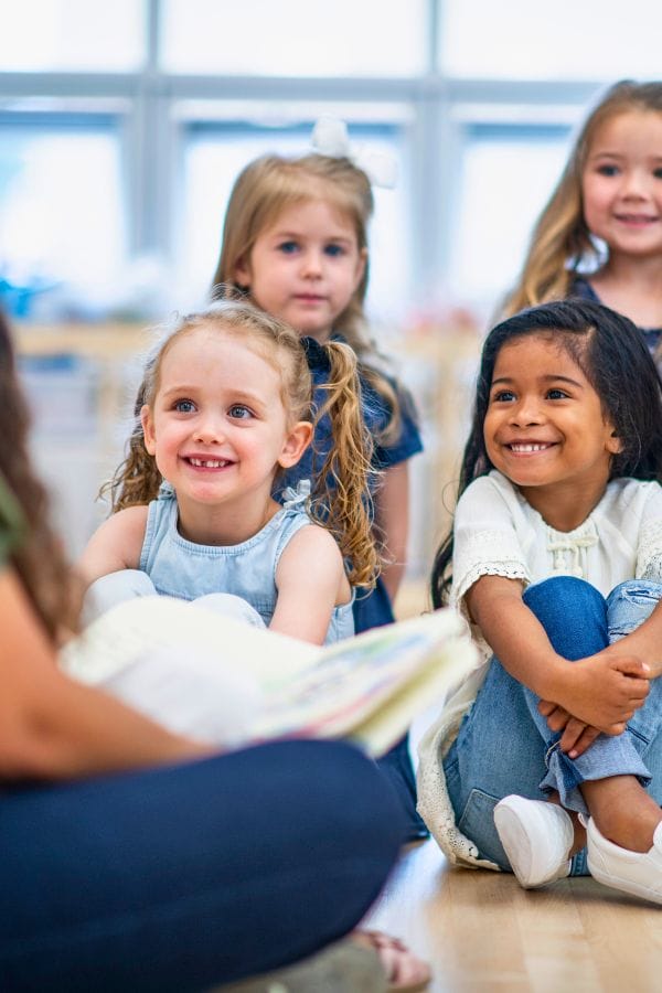 Children sit on the floor, smiling attentively as they engage in a daycare lesson on Pete the Cat. A woman, slightly out of focus in the foreground, hints at an exciting group activity or story time unfolding.