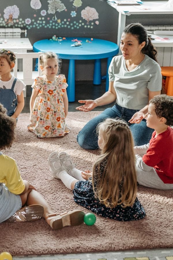 A woman sits on the floor with a group of young children, engaged in conversation about Pete the Cat. The children, seated in a semicircle, eagerly listen as toys related to their daycare lesson plans are scattered on the rug and nearby tables.