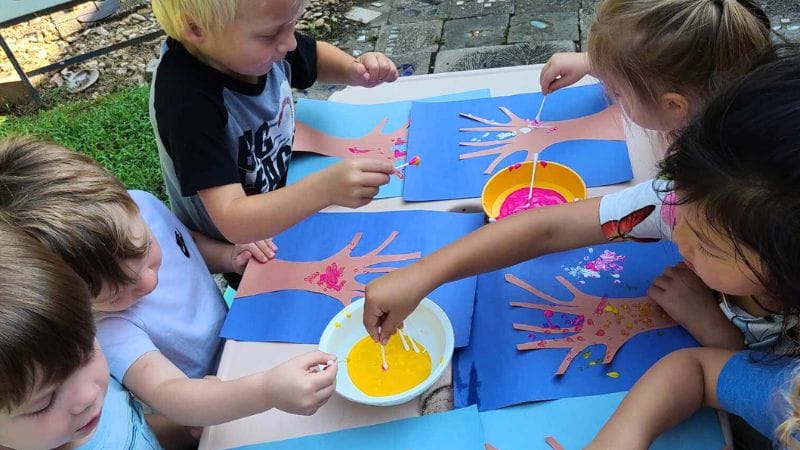 Children are seated at a table, using cotton swabs to create pointillism handprint trees with bright colors outdoors.