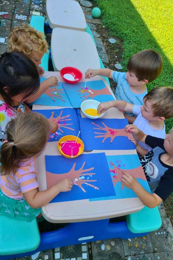 Children sitting at a table are eagerly engaged in a creative project, blending handprint art with the intricate style of pointillism, using paper and paint to create vibrant dot paintings.