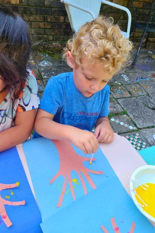 A child with curly hair engages in pointillism, dot painting a paper cut-out of a hand placed on blue paper. Another child is partially visible to the left, as they enjoy this outdoor activity on a mosaic-tiled surface.