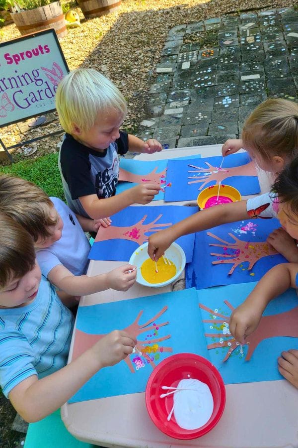 Children gather around a table outside, using cotton swabs to explore pointillism on blue paper. Bowls of paint and glue sit nearby, inspiring dot painting creativity.