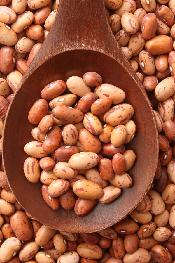 A wooden spoon filled with pinto beans rests on a surface covered with more pinto beans, inspiring you to grow pinto beans at home for fresh and hearty meals.