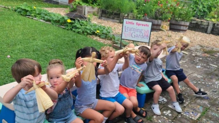 Children in the garden eagerly hold up their handcrafted corn husk dolls. A sign reads "Little Sprouts Learning Garden," surrounded by lush greenery and a charming stone path underfoot.