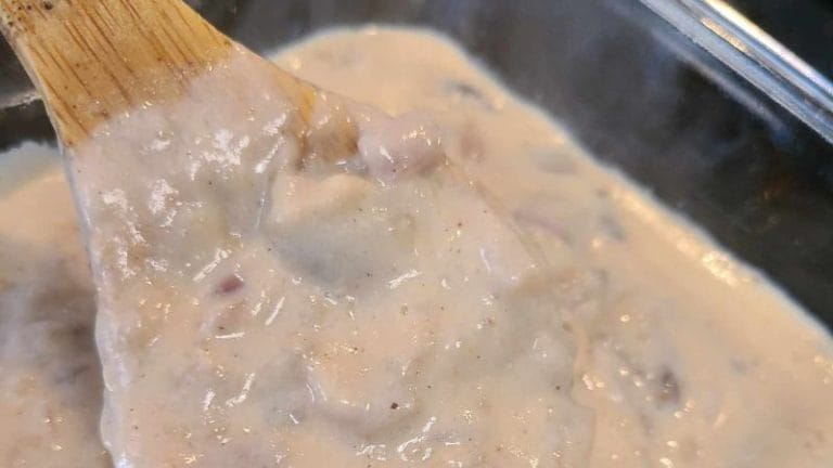 Close-up of a wooden spoon stirring a creamy, textured beige mixture with small visible bits—reminiscent of a comforting cream of anything soup—inside a transparent container.