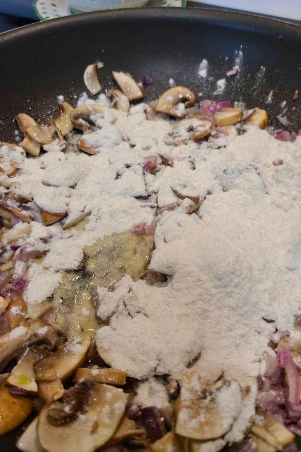 Mushrooms and chopped onions are being sautéed in a pan, ready to be transformed into a creamy base, as they're topped with a pile of flour—the ideal start for a comforting Cream of Anything Soup.