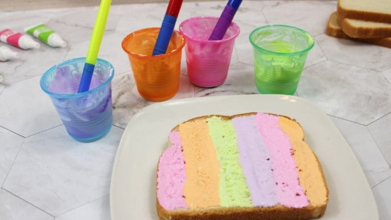 A slice of bread with pastel-colored stripes sits beside cups of edible paint, complete with brushes for artistic culinary creations.