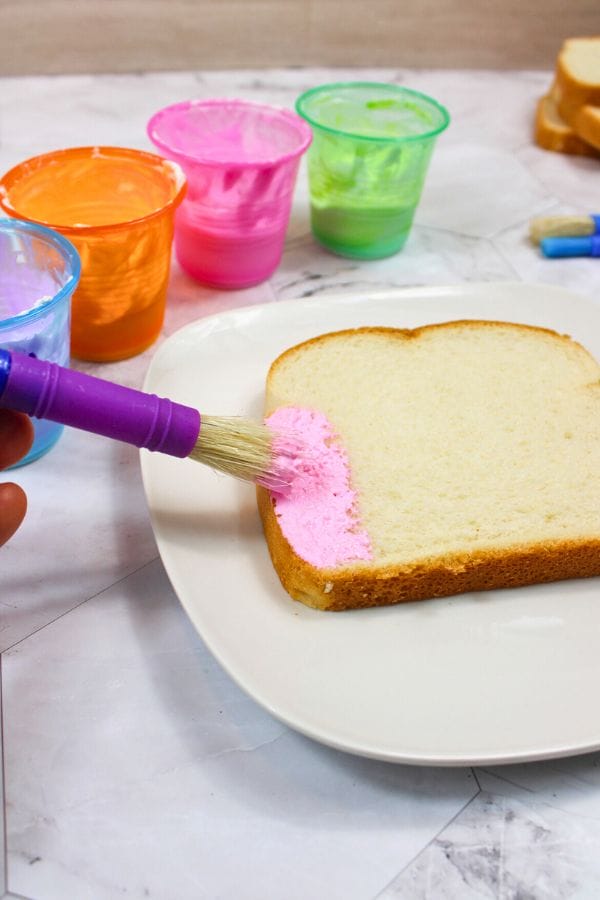 A slice of white bread on a plate is being painted with pink edible paint using a brush. Containers of various vibrant edible colors are in the background, ready to inspire culinary creativity.