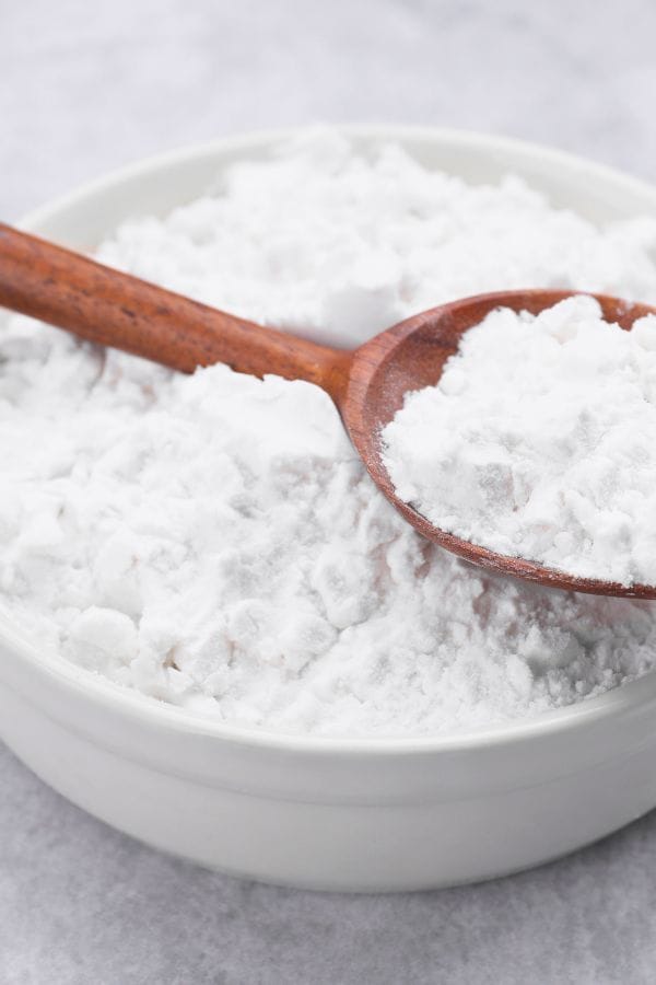 A wooden spoon rests on a bowl filled with white baking powder, ready to transform it into edible paint on a gray surface.