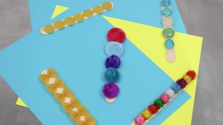 Five tactile sensory sticks decorated with various textured materials, including pom-poms, beads, sequins, and felt, are arranged on overlapping blue and yellow paper.