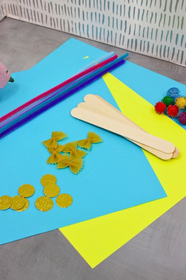 Craft materials including colored paper, tactile sensory sticks, pipe cleaners, glitter circles, uncooked pasta, colorful pom-poms, and a glue gun on a table await your creativity.