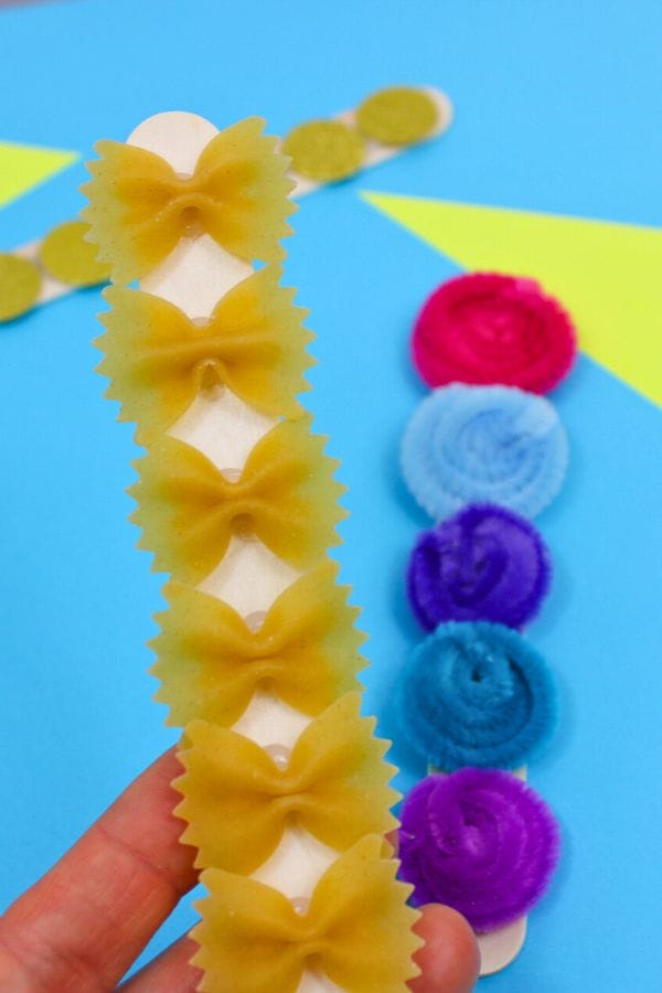 A hand holds a Tactile Sensory Stick adorned with bowtie pasta, while in the background, another stick featuring pom-poms rests on a blue surface dotted with yellow shapes.