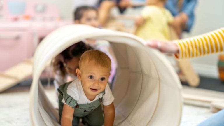 In a small home daycare, a baby crawls out of a fabric tunnel with a cheerful expression, surrounded by other children in the playroom.