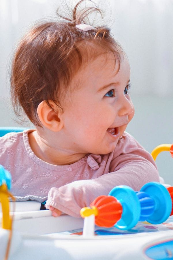 A baby with brown hair, wearing a pink shirt, is smiling and sitting in a colorful play activity center at a small home daycare filled with various toys.