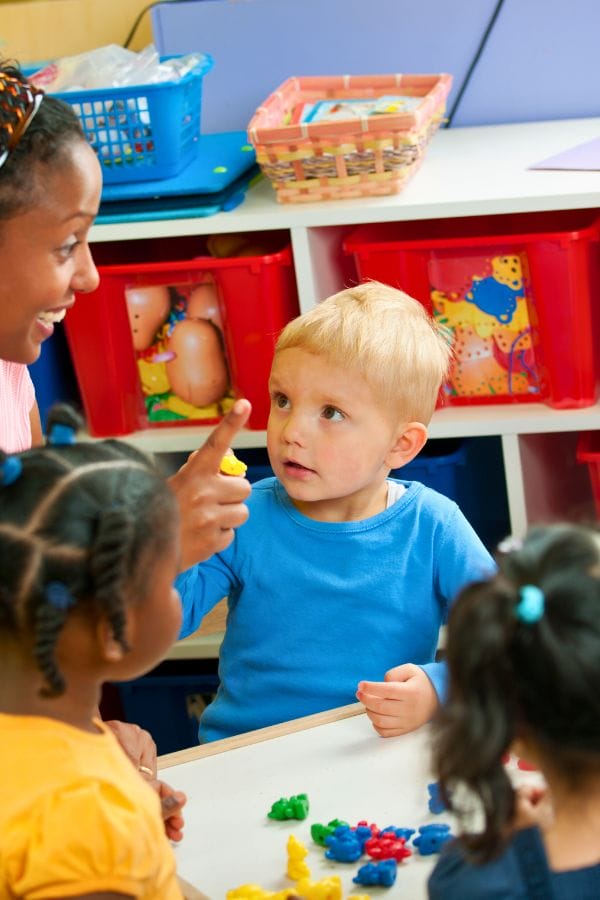 In a cozy small home daycare, a young child in a blue shirt points excitedly while seated at a table filled with colorful toys, engaging with a woman. Nearby, two other children are deeply focused on the lively activities around them.
