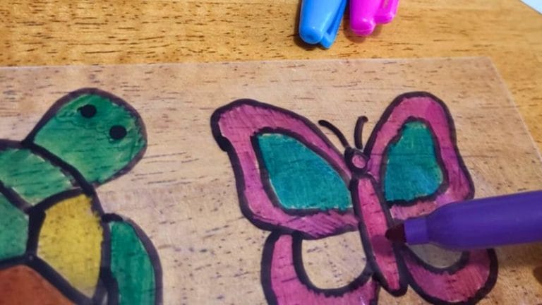 A close-up of a hand coloring a butterfly drawing with a purple marker next to a colored turtle drawing on a wooden surface; blue and pink markers are nearby, perfect for creating Summer Shrinky Dink Patterns.