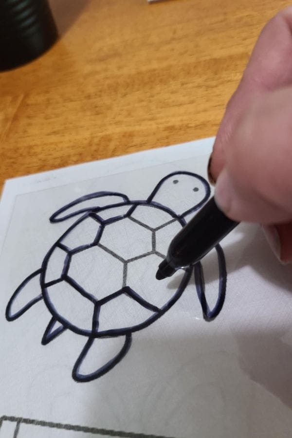 A hand uses a black marker to trace the outline of a turtle drawing on a piece of paper, creating one of the Summer Shrinky Dink Patterns on a wooden surface.