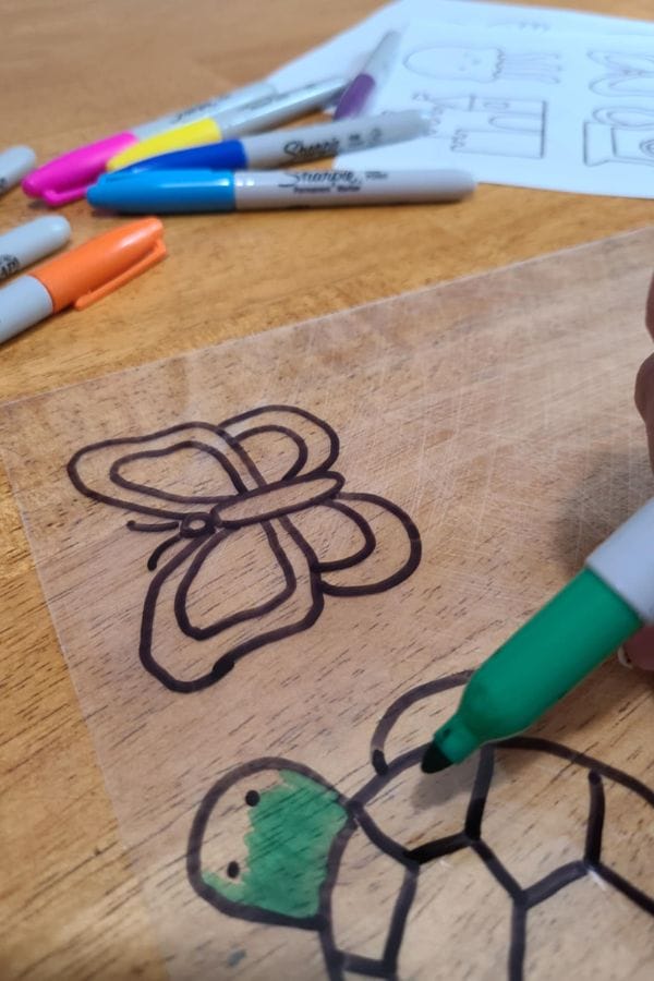 A hand colors a turtle drawing on a clear plastic sheet with a green marker; Summer Shrinky Dink Patterns, like the butterfly seen nearby, and markers are also visible on the table.