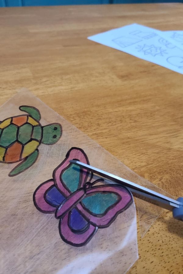 A hand cuts out colored drawings of a turtle and butterfly from a clear sheet on a wooden table, using Summer Shrinky Dink Patterns, with more vibrant designs visible in the background.