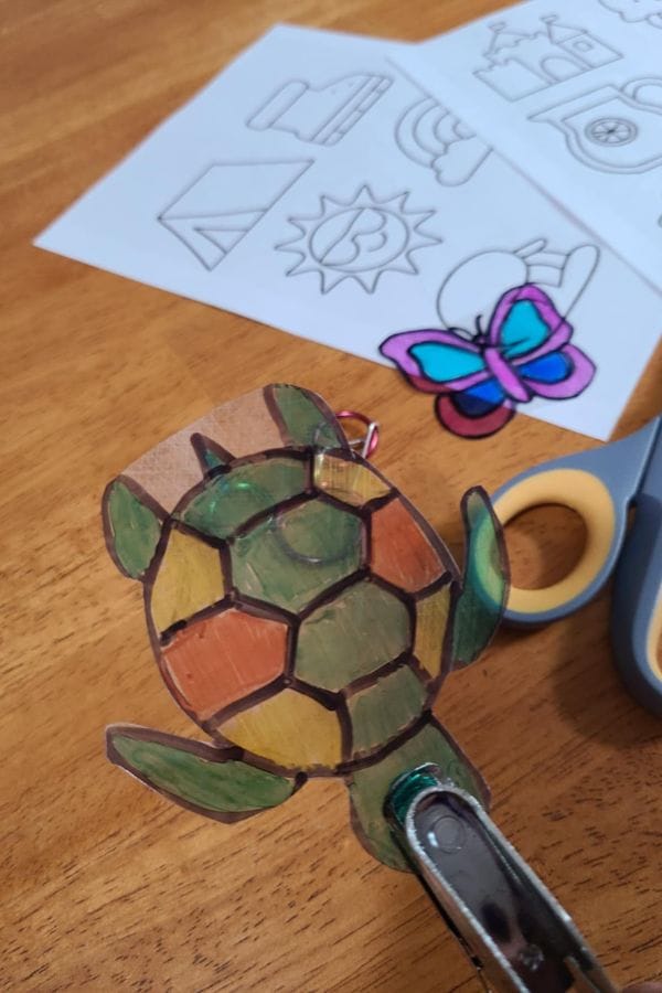 A colored turtle and butterfly drawing on shrink plastic, with scissors and uncolored templates on a wooden table—perfect for creating Summer Shrinky Dink Patterns.