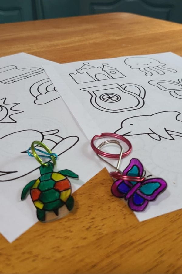 Two sheets of paper with outlined drawings, next to colorful Summer Shrinky Dink Patterns of turtle and butterfly keychains made from plastic and wire, on a wooden surface.