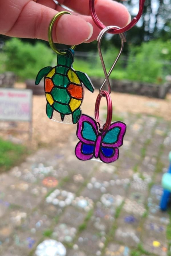 A hand holds two keychains made with Summer Shrinky Dink Patterns: one shaped like a multicolored turtle and the other like a purple butterfly, against an outdoor background with a stone path and greenery.