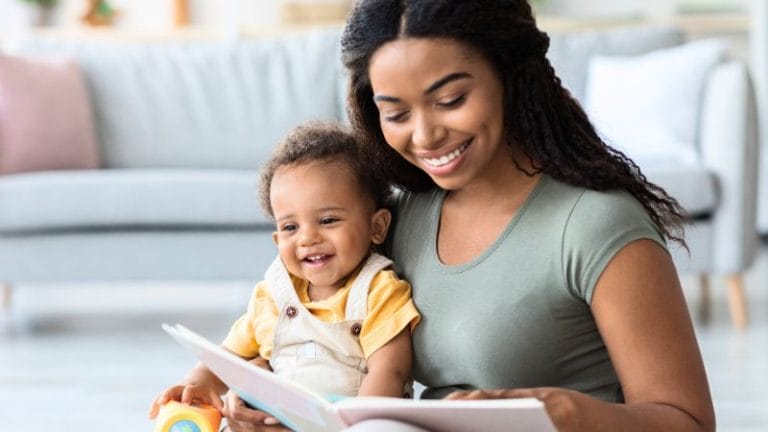 A woman reads a book to a smiling baby sitting on her lap in a living room with a couch in the background, showcasing one of the engaging daycare activities for infants.