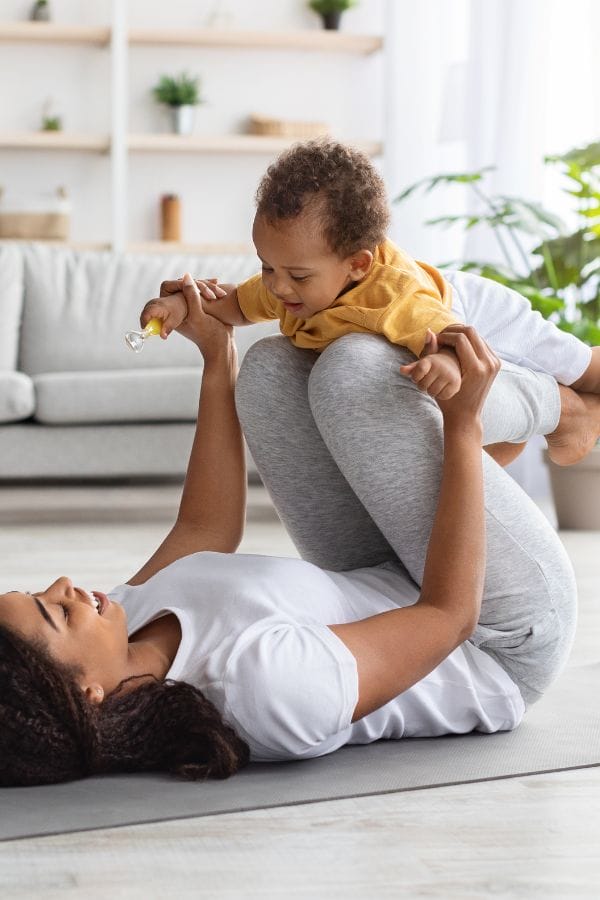 Woman lying on a mat holds a smiling toddler above her with bent knees in a living room, while the child holds a small toy—one of many fun daycare activities for infants that encourage bonding and development.
