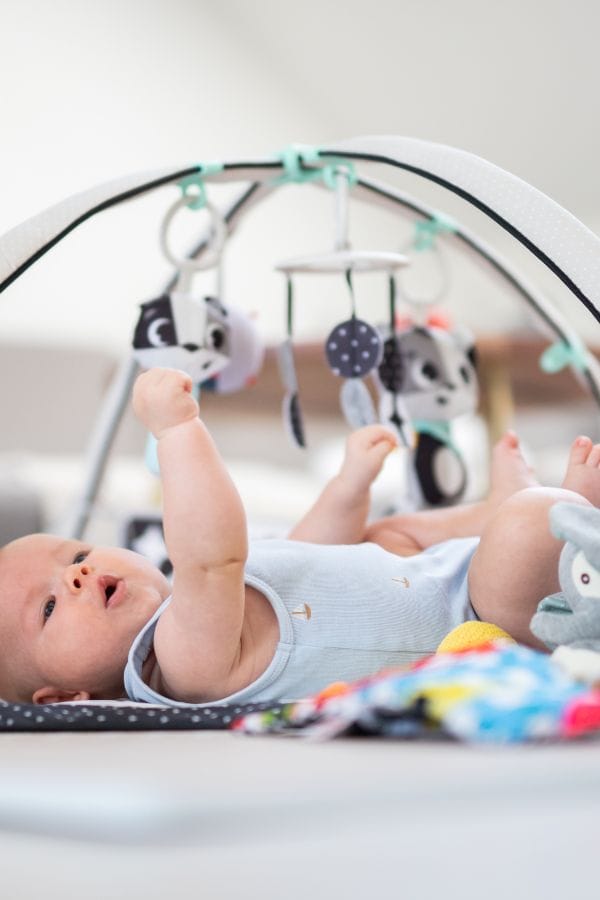 A baby lies on a play mat, reaching up towards hanging toys attached to an overhead play gym—one of the engaging daycare activities for infants.