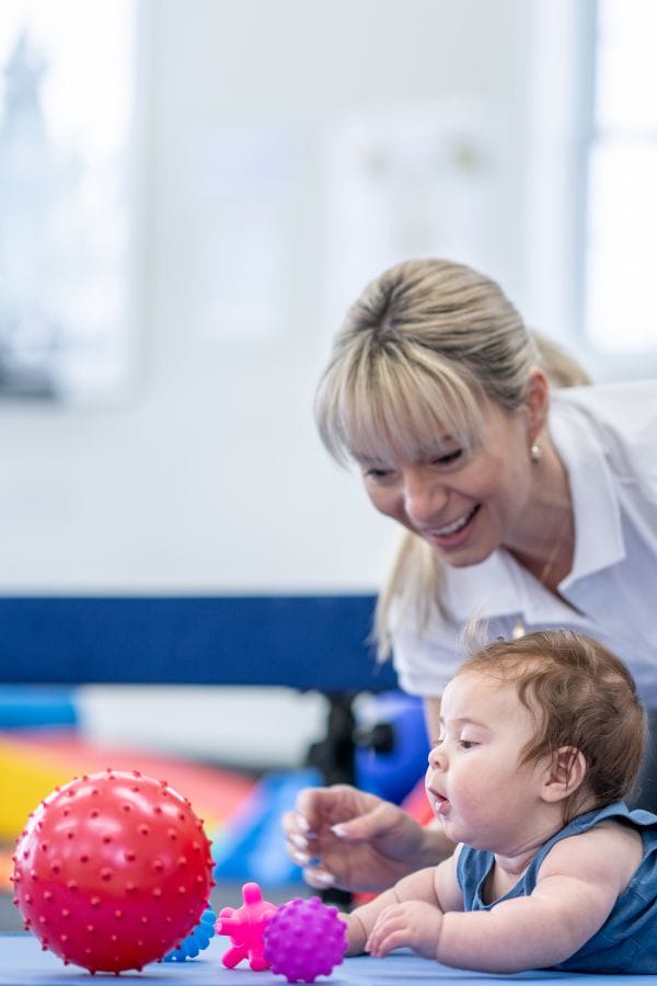 A woman smiles as she assists a baby who is lying on their stomach and reaching toward colorful textured balls on a mat, engaging in fun daycare activities for infants.